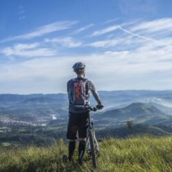 A cyclist stands on a hill, gazing at a stunning mountain landscape on a bright sunny day.