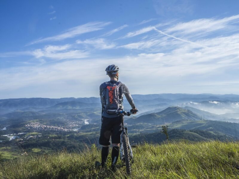 A cyclist stands on a hill, gazing at a stunning mountain landscape on a bright sunny day.