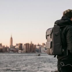A man wearing a backpack gazes at the Manhattan skyline from Brooklyn waterfront during the day.