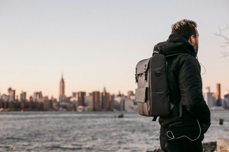 A man wearing a backpack gazes at the Manhattan skyline from Brooklyn waterfront during the day.