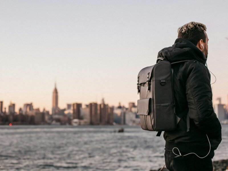 A man wearing a backpack gazes at the Manhattan skyline from Brooklyn waterfront during the day.